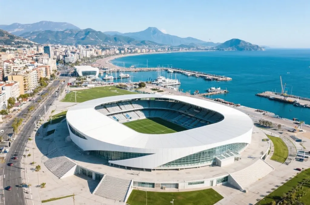 An aerial view of a modern soccer stadium near a scenic coastal city harbor with distant mountains, daytime, clean architectural design